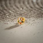 A gold round-frame stud earring set with multicolor stones in purple, pink, green, red and white, placed on a textured beige fabric background