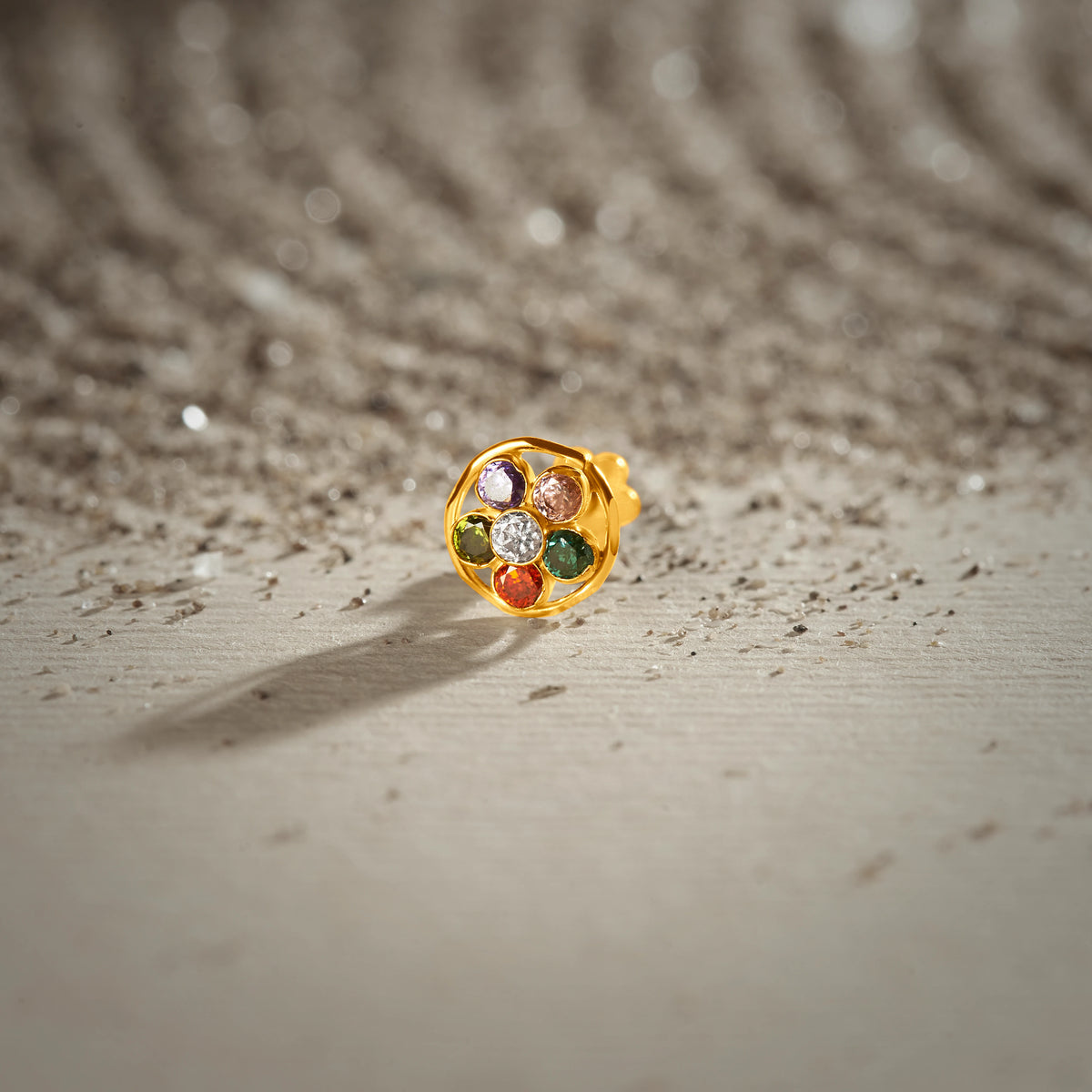 A gold round-frame stud earring set with multicolor stones in purple, pink, green, red and white, placed on a textured beige fabric background