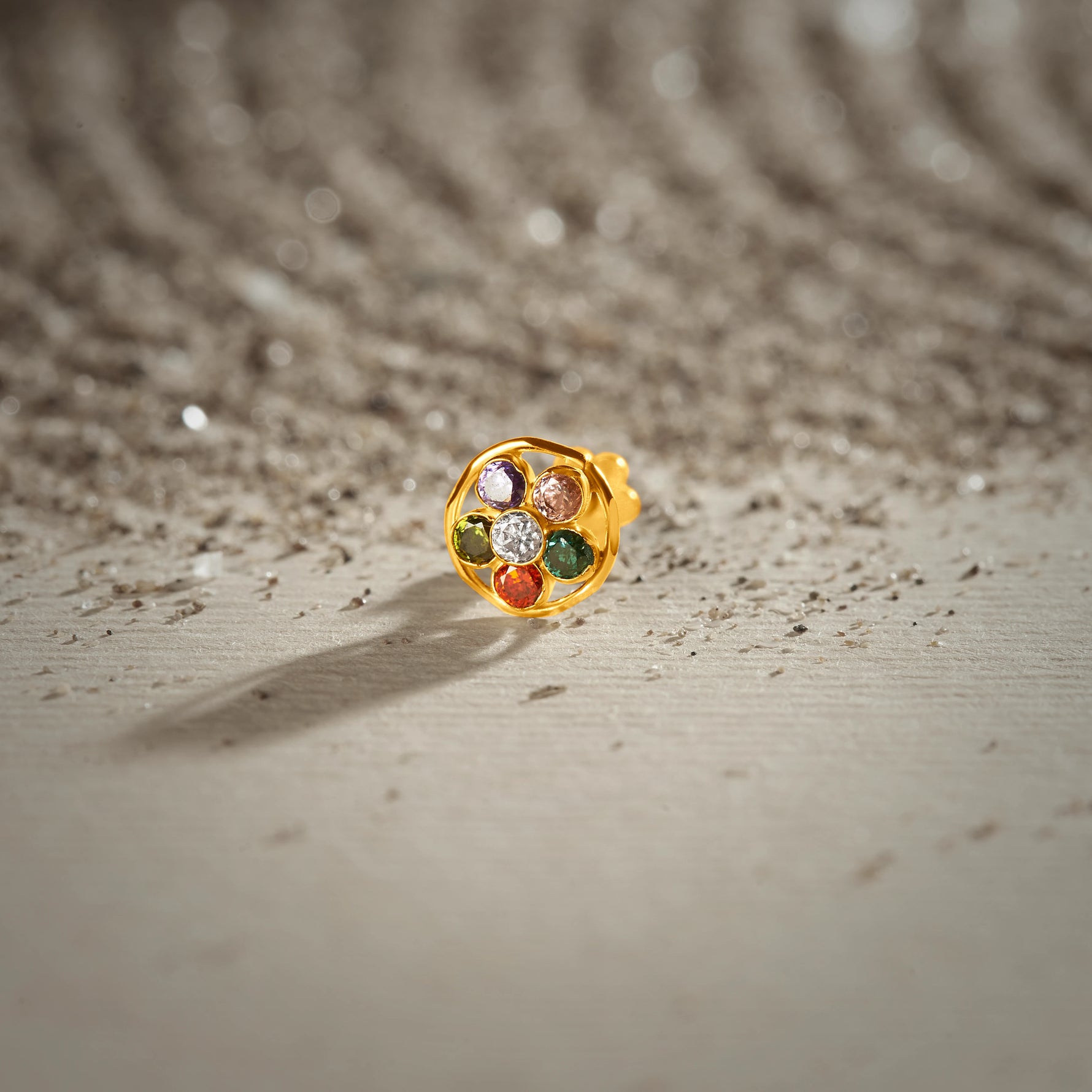 A gold round-frame stud earring set with multicolor stones in purple, pink, green, red and white, placed on a textured beige fabric background