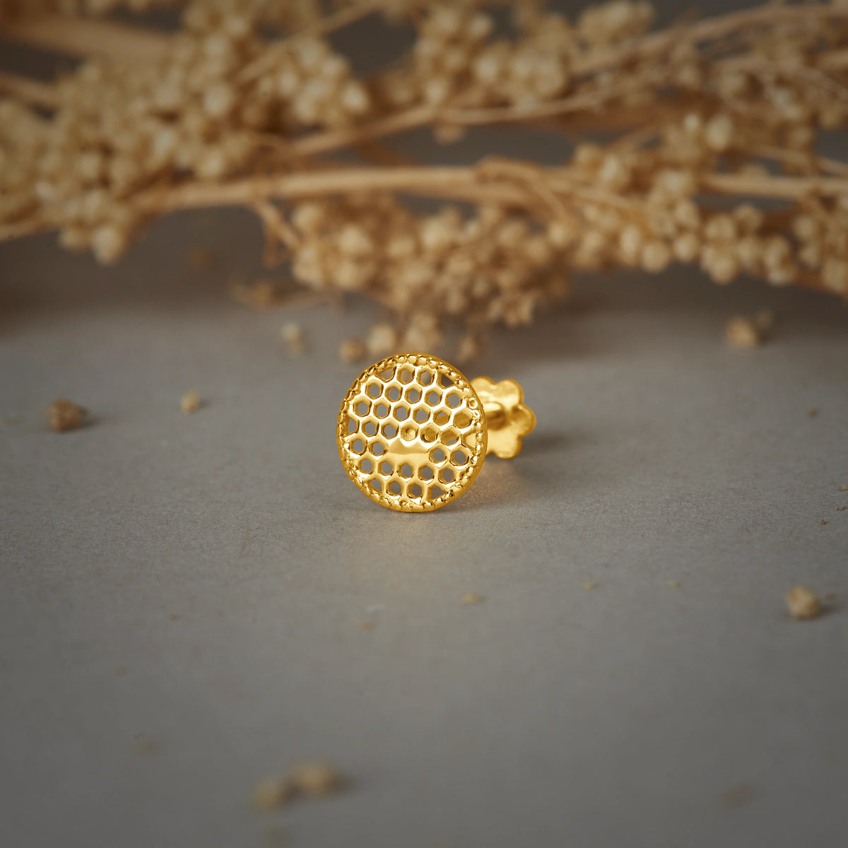 A round gold stud earring with a honeycomb cutwork pattern, placed on a grey background with dried flower branches
