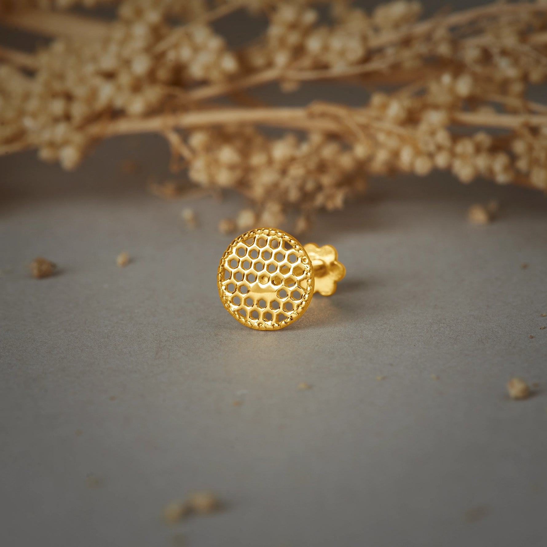 A round gold stud earring with a honeycomb cutwork pattern, placed on a grey background with dried flower branches