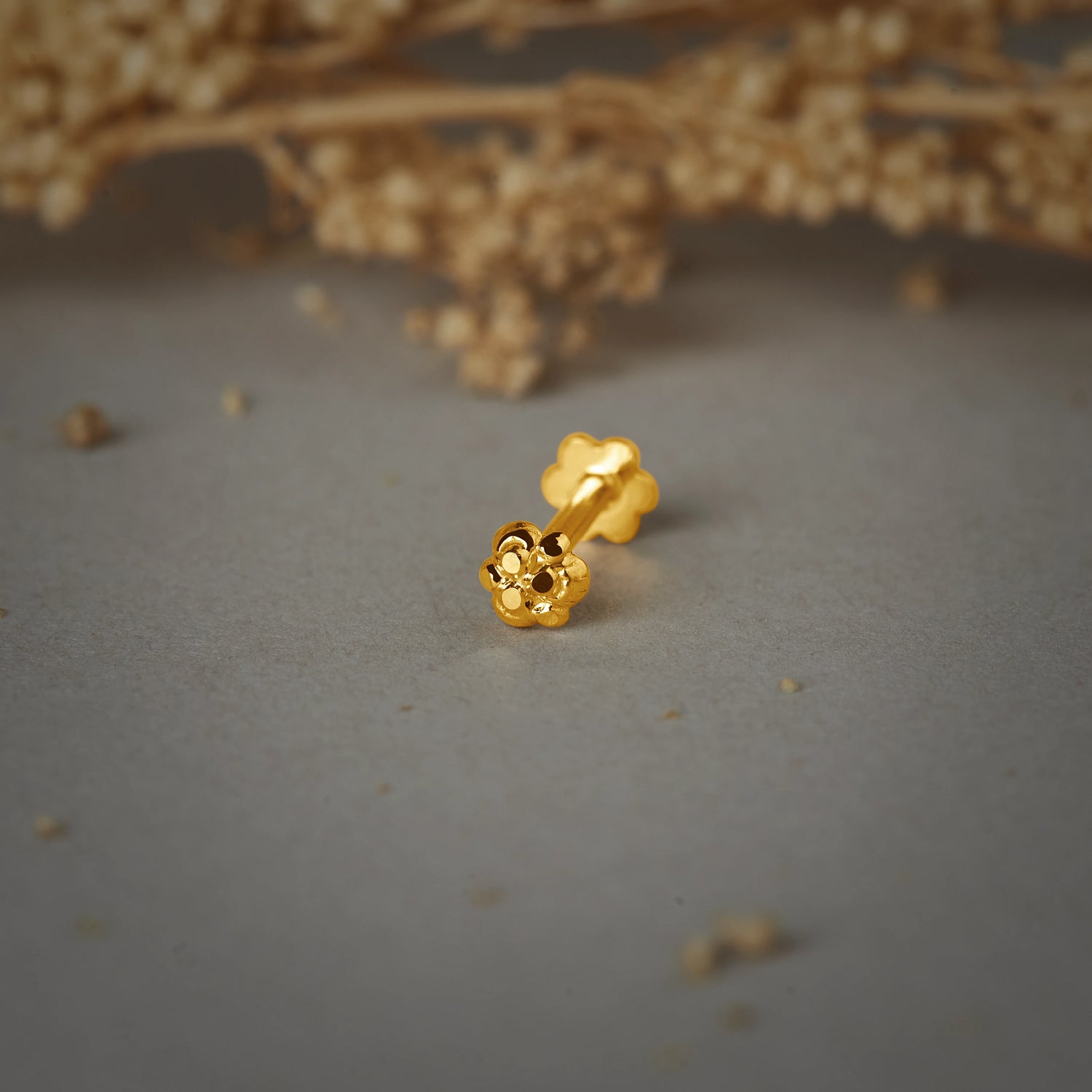 A gold flower-shaped stud earring with rounded petals and a beaded center, placed on a grey background with dried flowers
