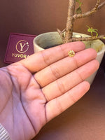 Small round textured gold nose pin held on fingers for size comparison, with Yuvora jewellery box and plant in background