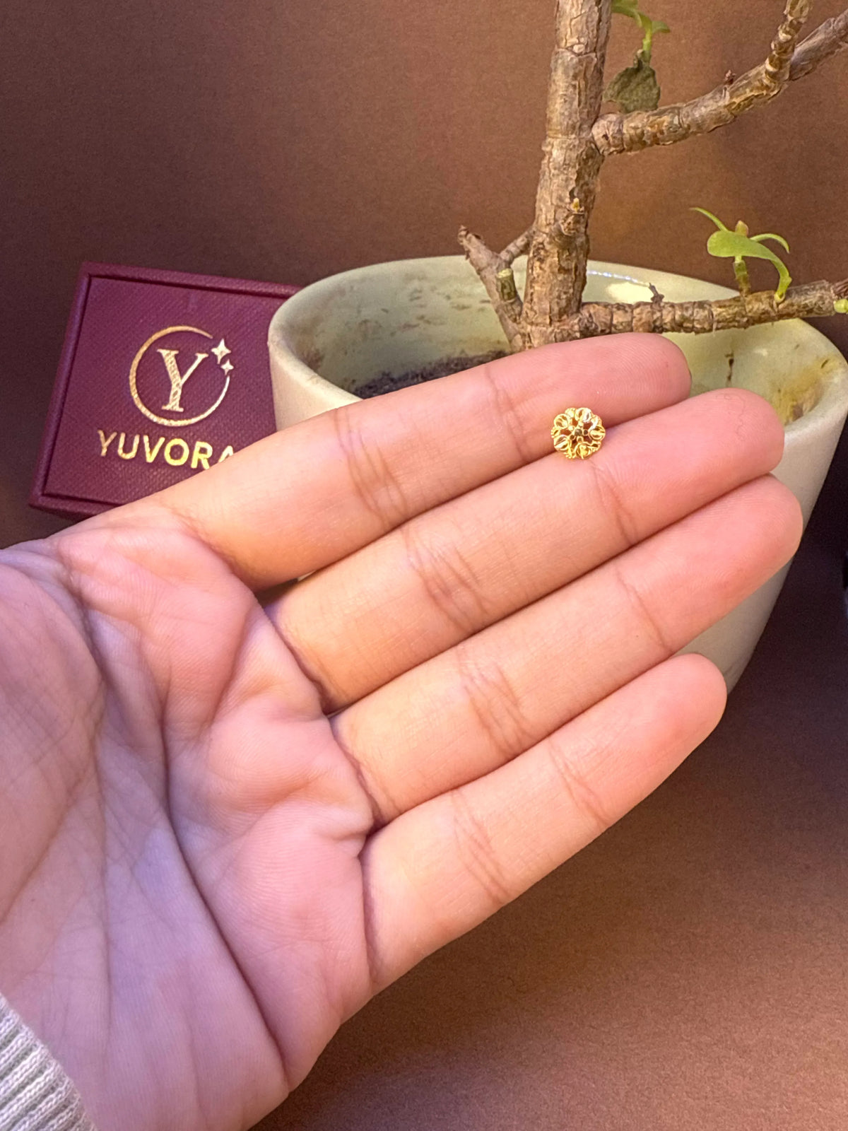 Small round textured gold nose pin held on fingers for size comparison, with Yuvora jewellery box and plant in background