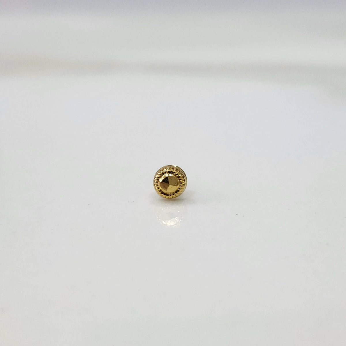 Round gold nose pin with a faceted center and textured border on a white surface