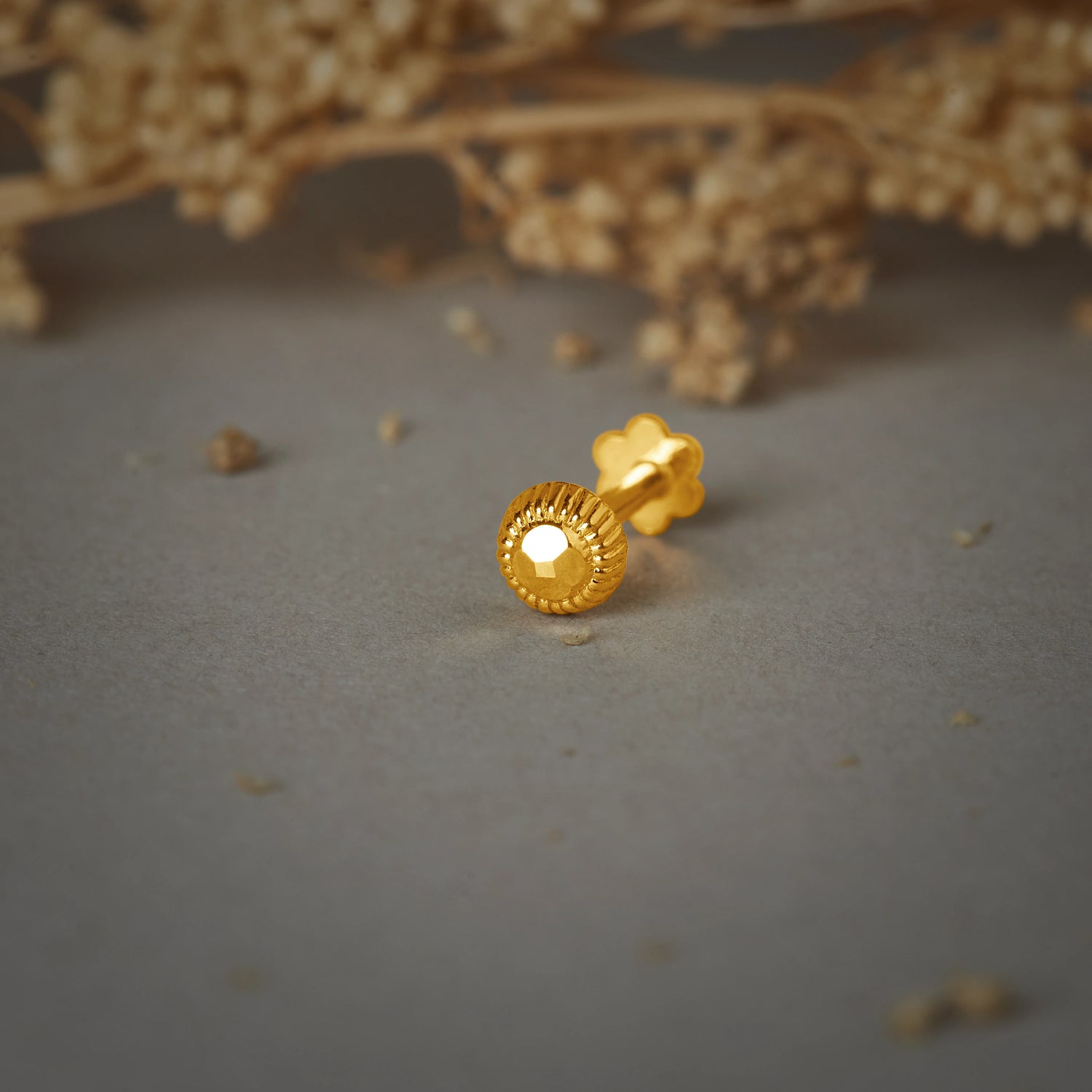 A round gold nose pin with a ribbed edge and a faceted center, placed on a grey background with dried flowers