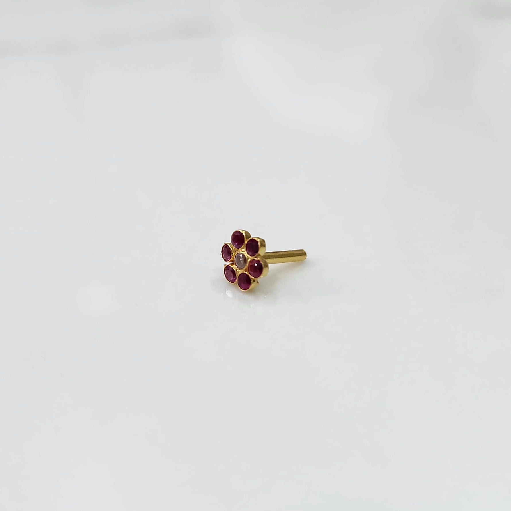 A gold flower-shaped nose pin with ruby stones around a small champagne-colored center stone, placed on a white background