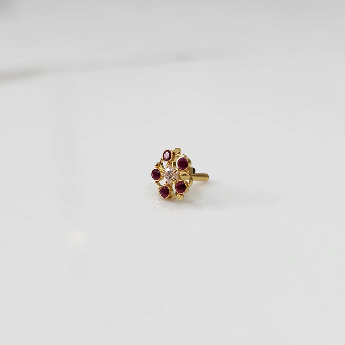 Gold nose pin with ruby-colored gemstone petals and clear center stone on white background