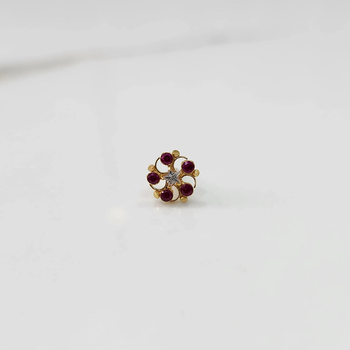 Front view of gold nose pin with ruby-colored gemstone petals and clear center stone on white background 