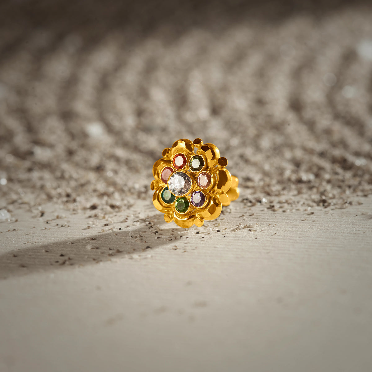 A gold scalloped-edge stud earring with a white center stone surrounded by multicolor stones in red, green, purple and cream, placed on a textured beige fabric background