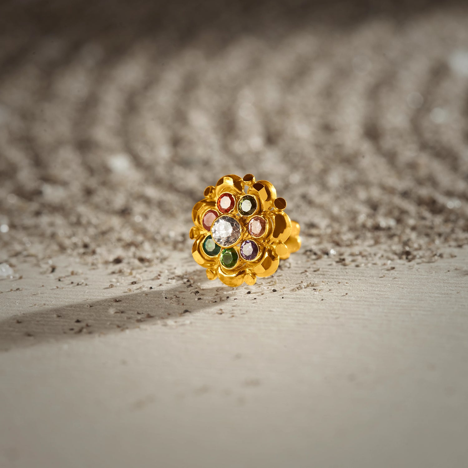 A gold scalloped-edge stud earring with a white center stone surrounded by multicolor stones in red, green, purple and cream, placed on a textured beige fabric background