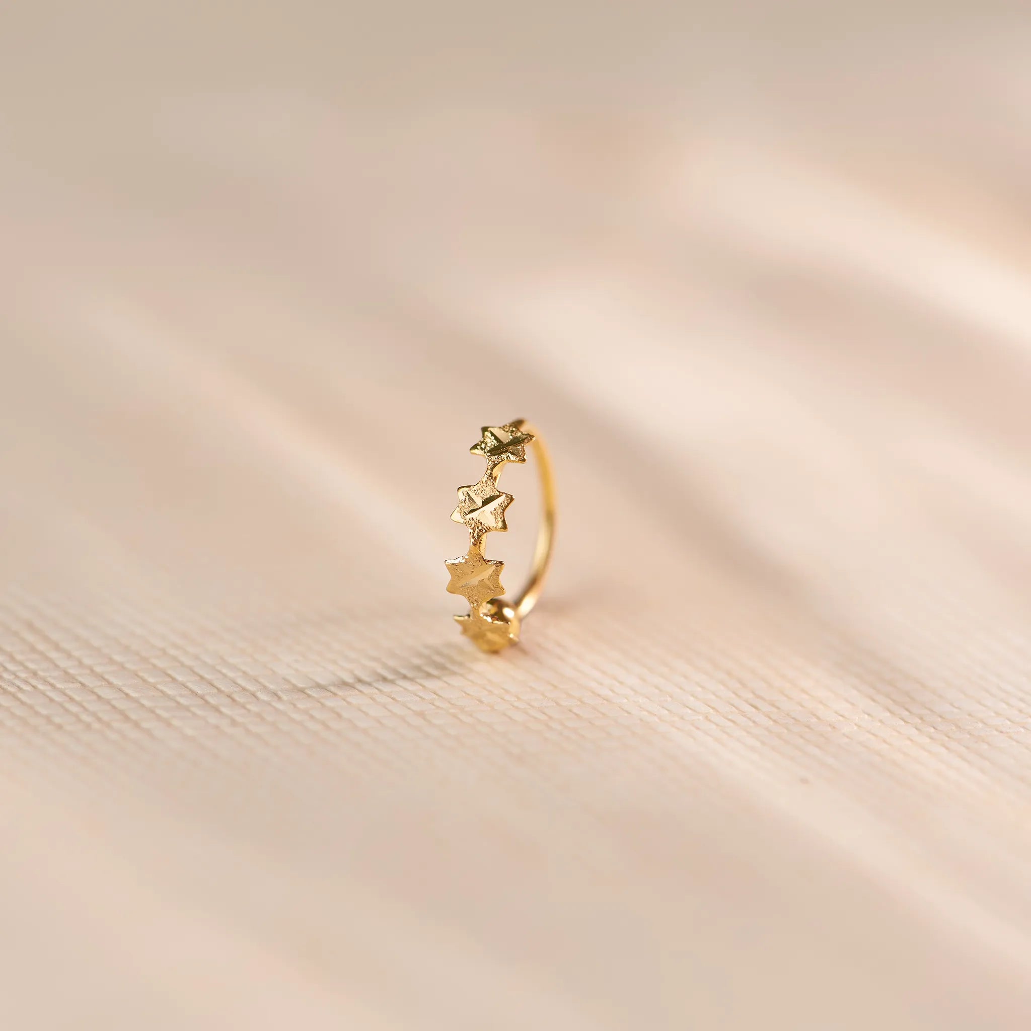 Close up of star patterned 18K gold nose ring standing upright on a soft beige fabric surface with gentle light shadows