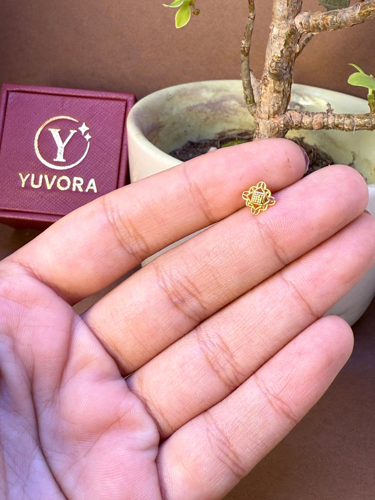 Small gold filigree star-flower nose pin held on fingers for size comparison, with Yuvora jewellery box and plant in background