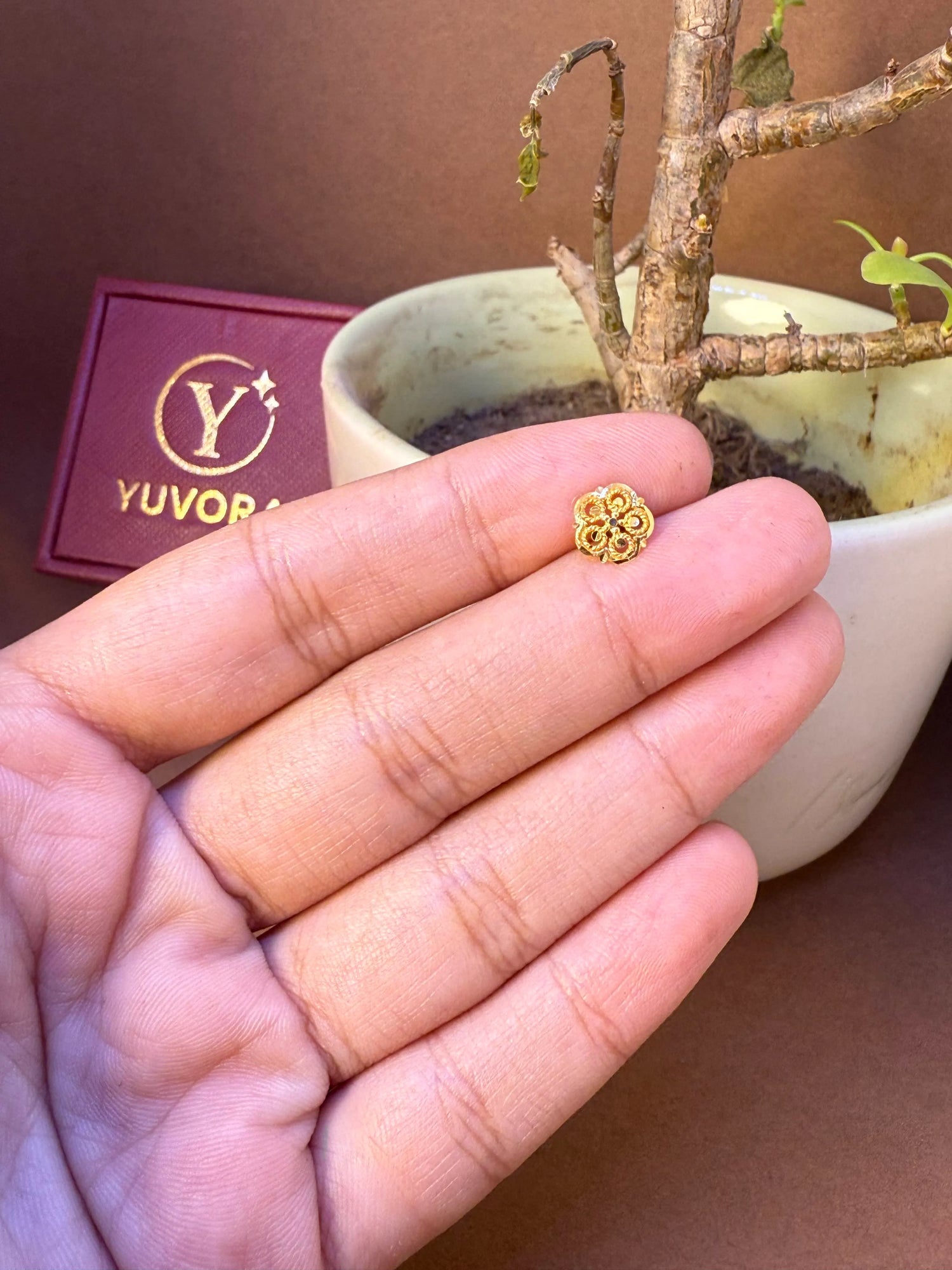 Small gold star flower nose pin held on fingers for size comparison, with Yuvora jewellery box and plant in background