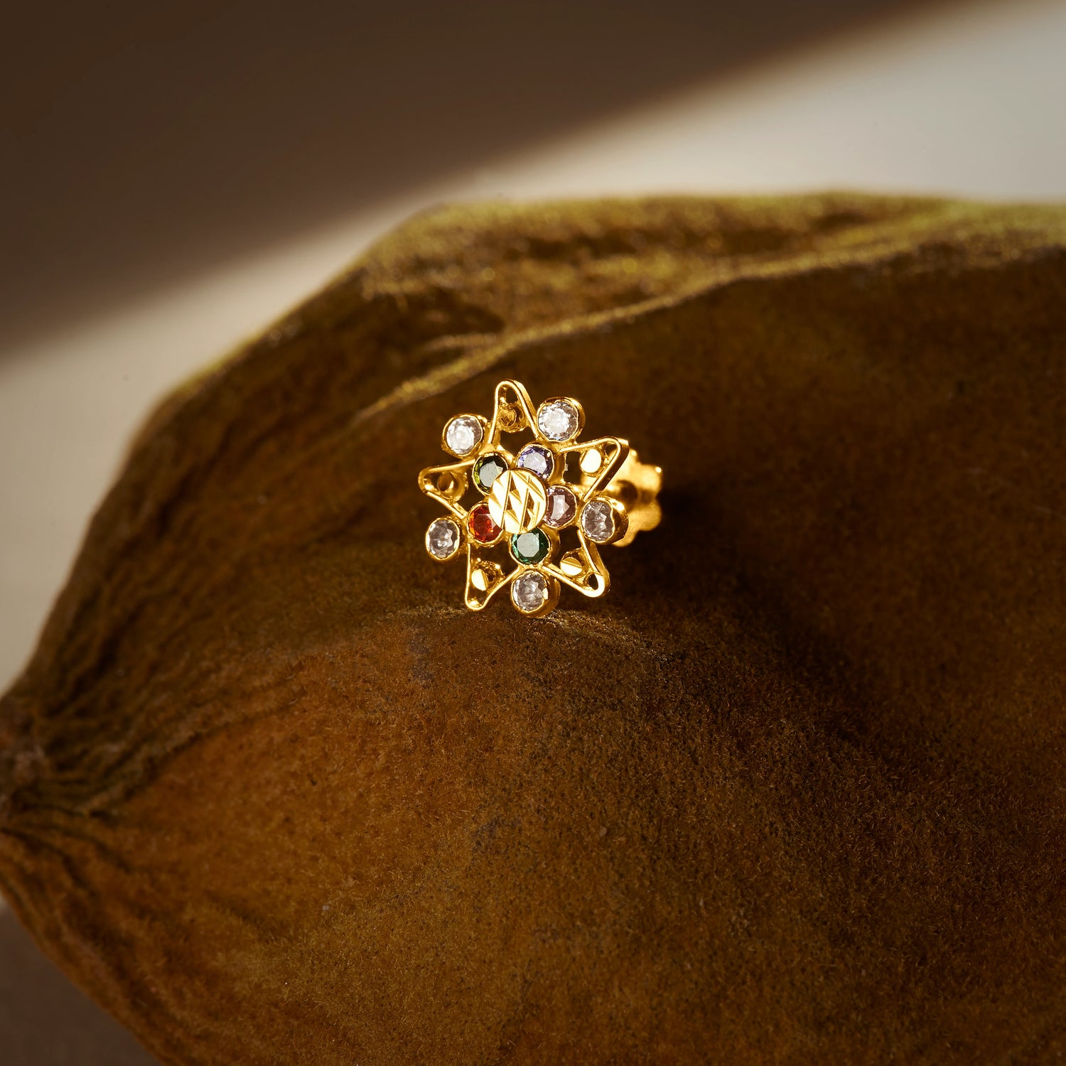 A gold star-shaped stud earring with a textured center bead and multicolor stones set along the star points, placed on a textured brown fabric background