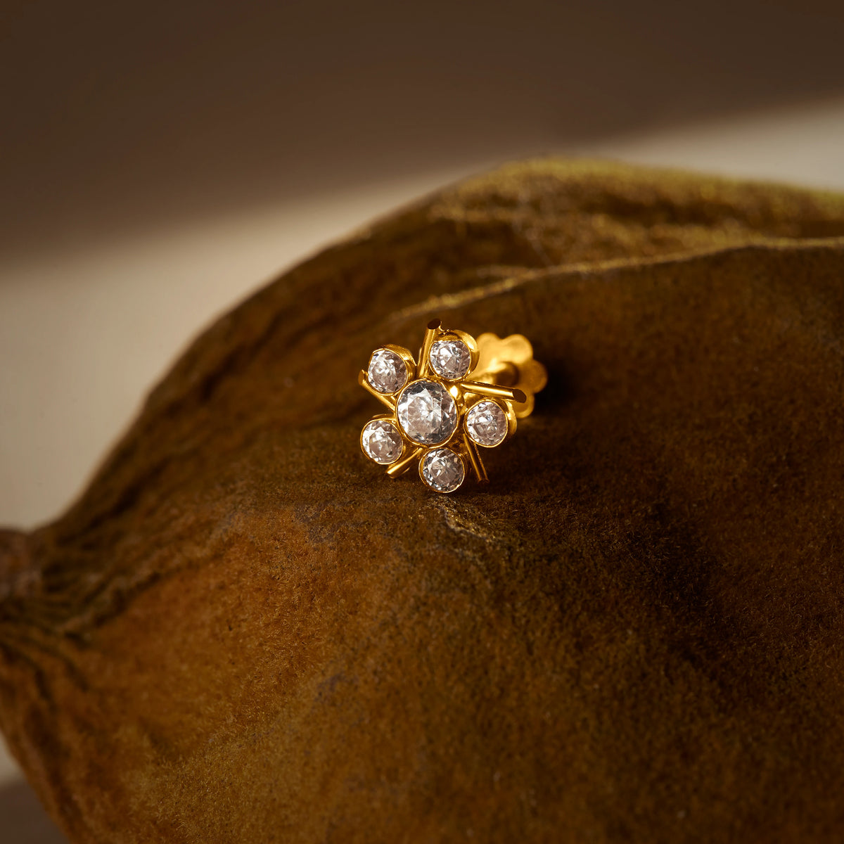 A gold flower-shaped stud earring with star-like prong settings holding five round white stones, placed on a textured brown fabric background