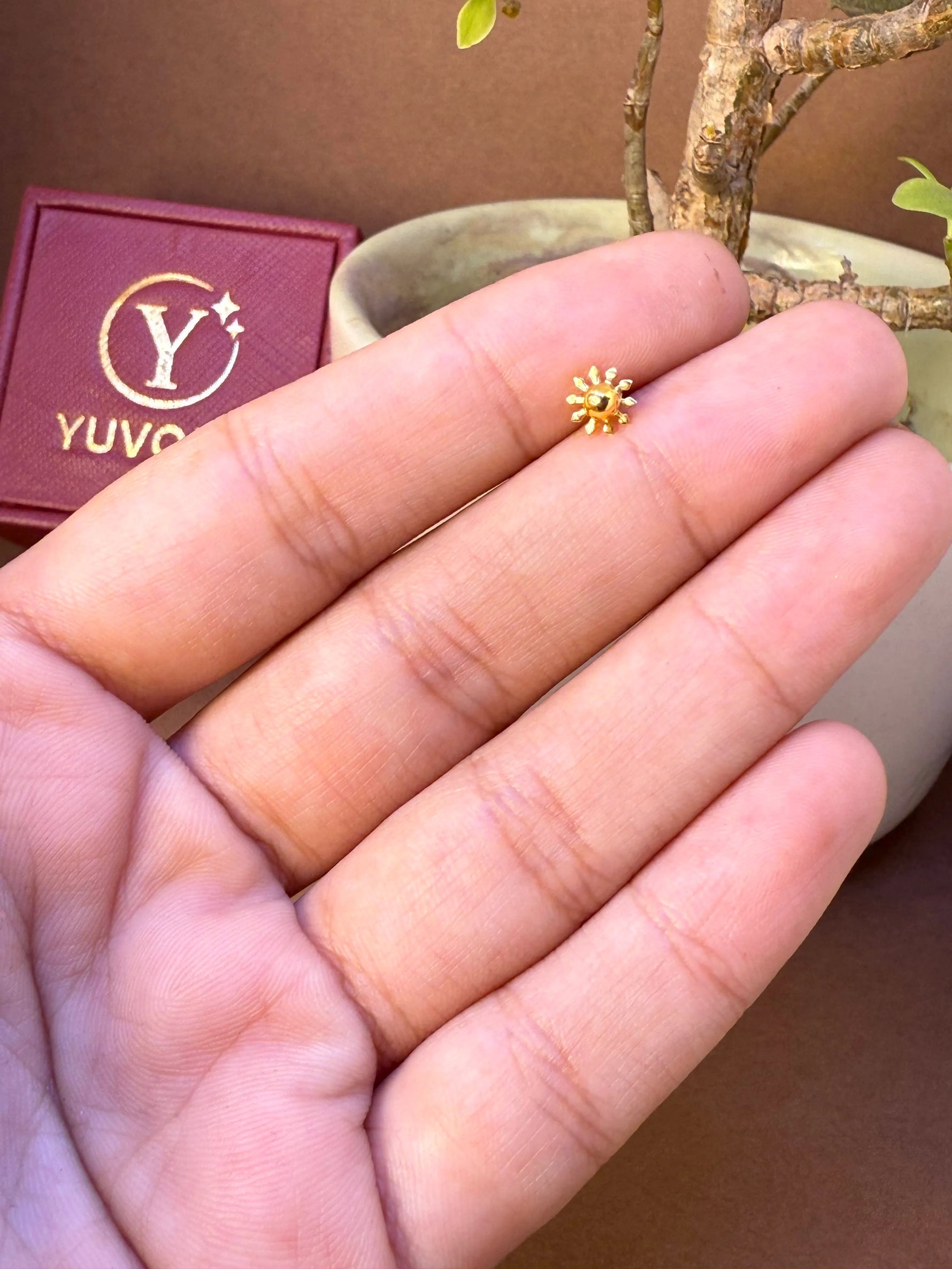 Small gold sun-shaped nose pin held on fingers for size comparison, with Yuvora jewellery box and plant in background