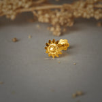 A gold sunflower-shaped nose pin with pointed petals and a textured center, placed on a grey background with dried flowers