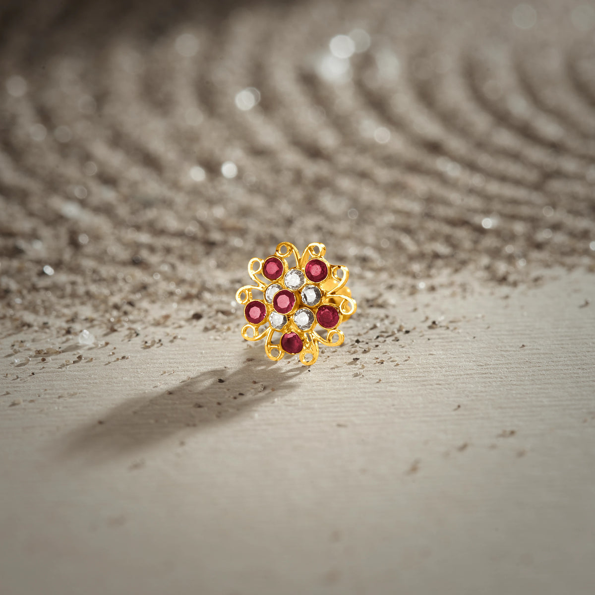 A gold swirl-design flower stud earring set with ruby and white stones, placed on a textured beige fabric background