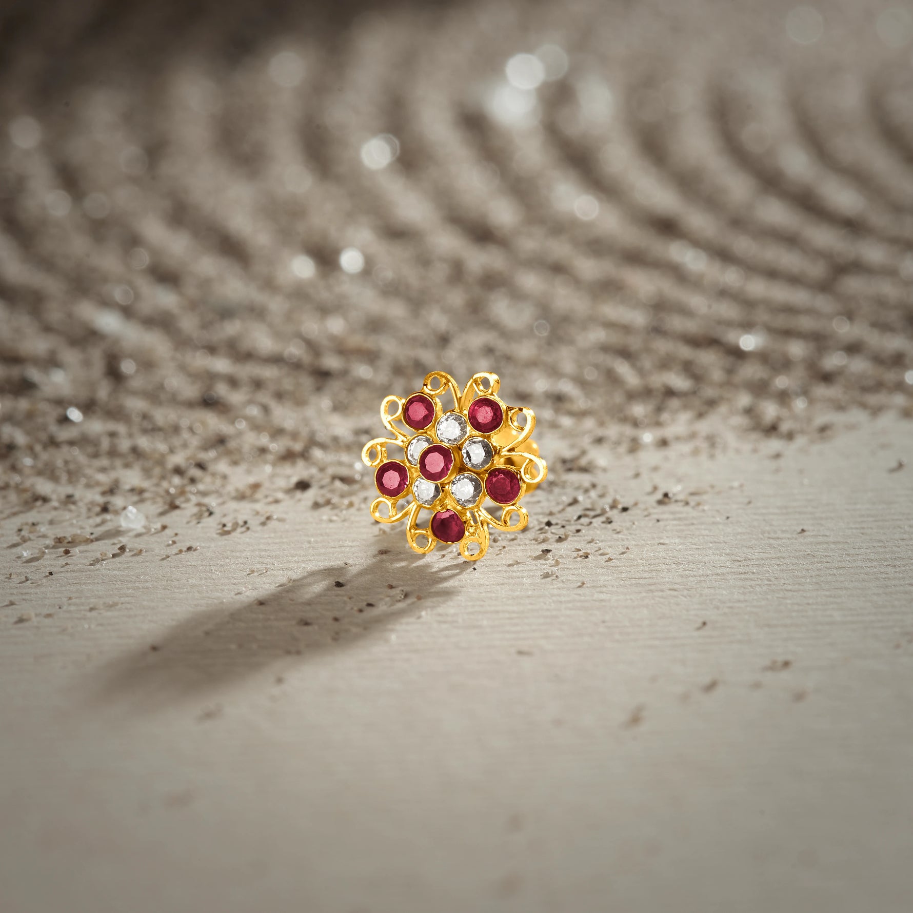 A gold swirl-design flower stud earring set with ruby and white stones, placed on a textured beige fabric background