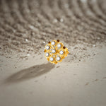 A gold swirl-petal flower stud earring with round white stones set in a beaded frame, placed on a textured beige fabric background