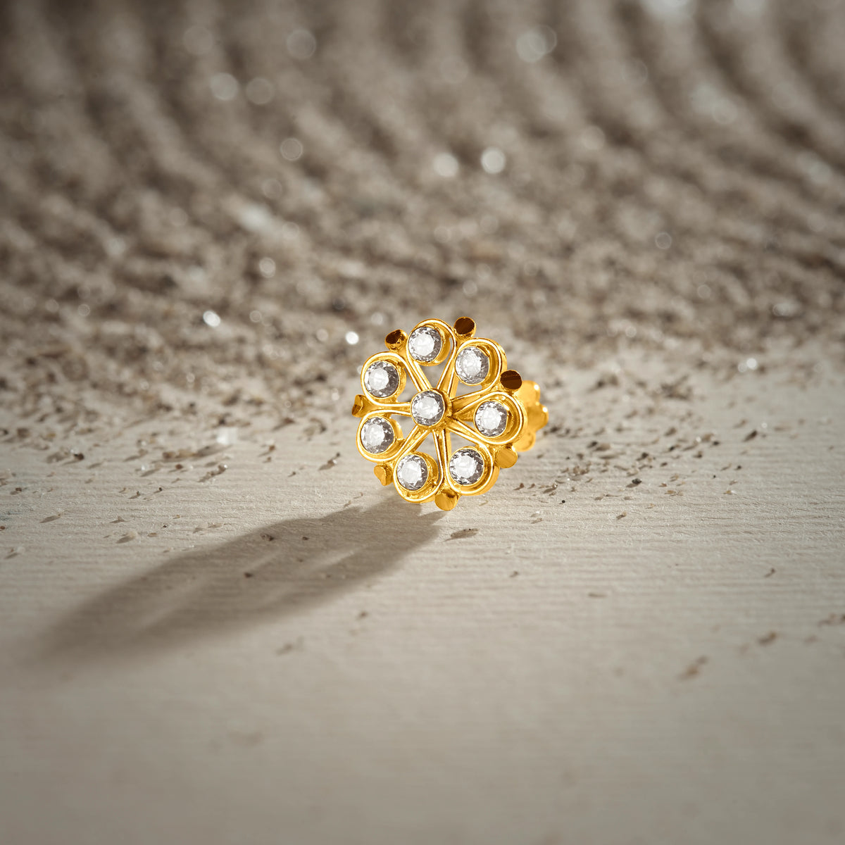 A gold swirl-petal flower stud earring with round white stones set in a beaded frame, placed on a textured beige fabric background