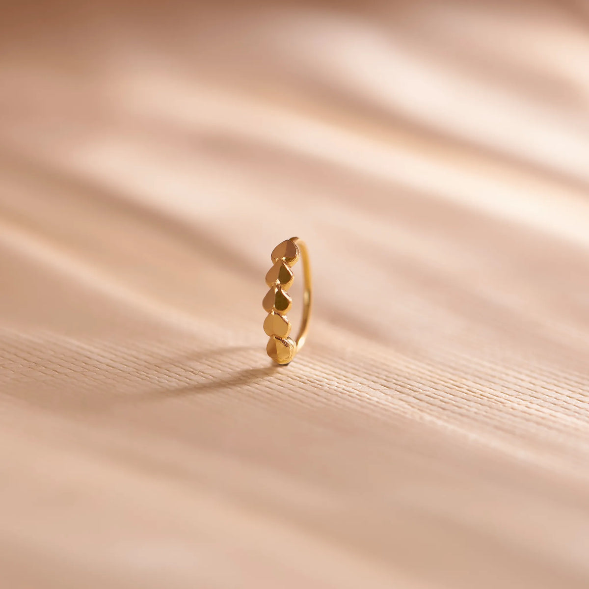 Close up of patterned 18K gold nose ring standing upright on a soft beige fabric background with flowing light textures