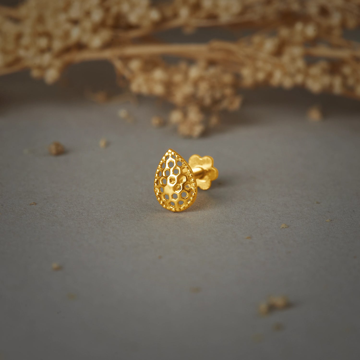 A gold teardrop-shaped stud earring with a honeycomb cutwork pattern, placed on a grey background with dried flowers