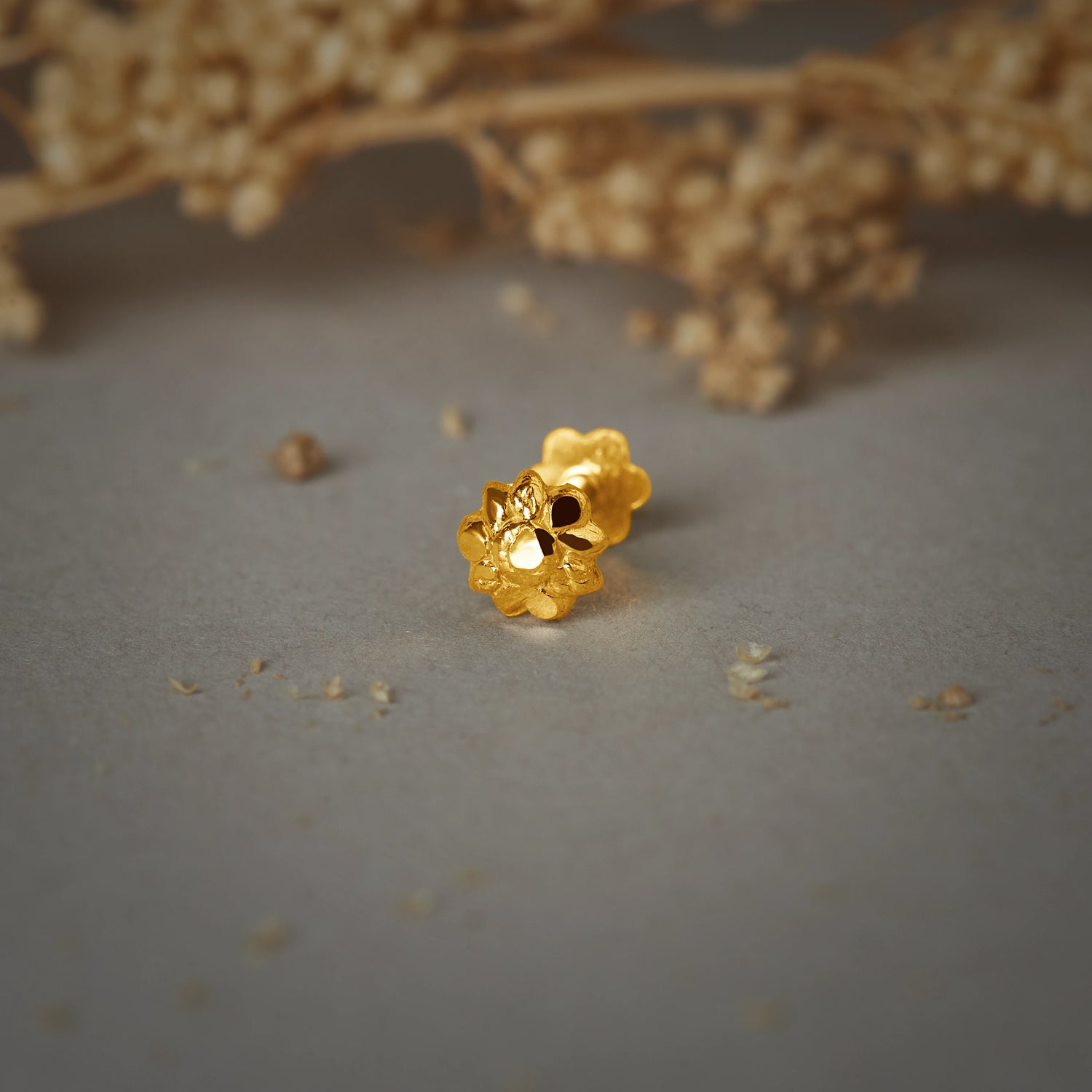 A gold flower-shaped stud earring with a textured petal design, placed on a grey background with dried flowers