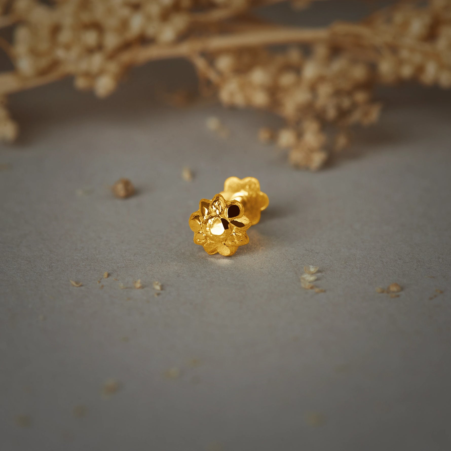 A gold flower-shaped stud earring with a textured petal design, placed on a grey background with dried flowers