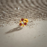 A gold trefoil-shaped stud earring with three ruby stones and a white center stone, placed on a textured beige fabric background