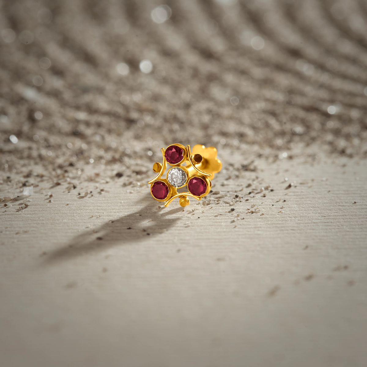 A gold trefoil-shaped stud earring with three ruby stones and a white center stone, placed on a textured beige fabric background