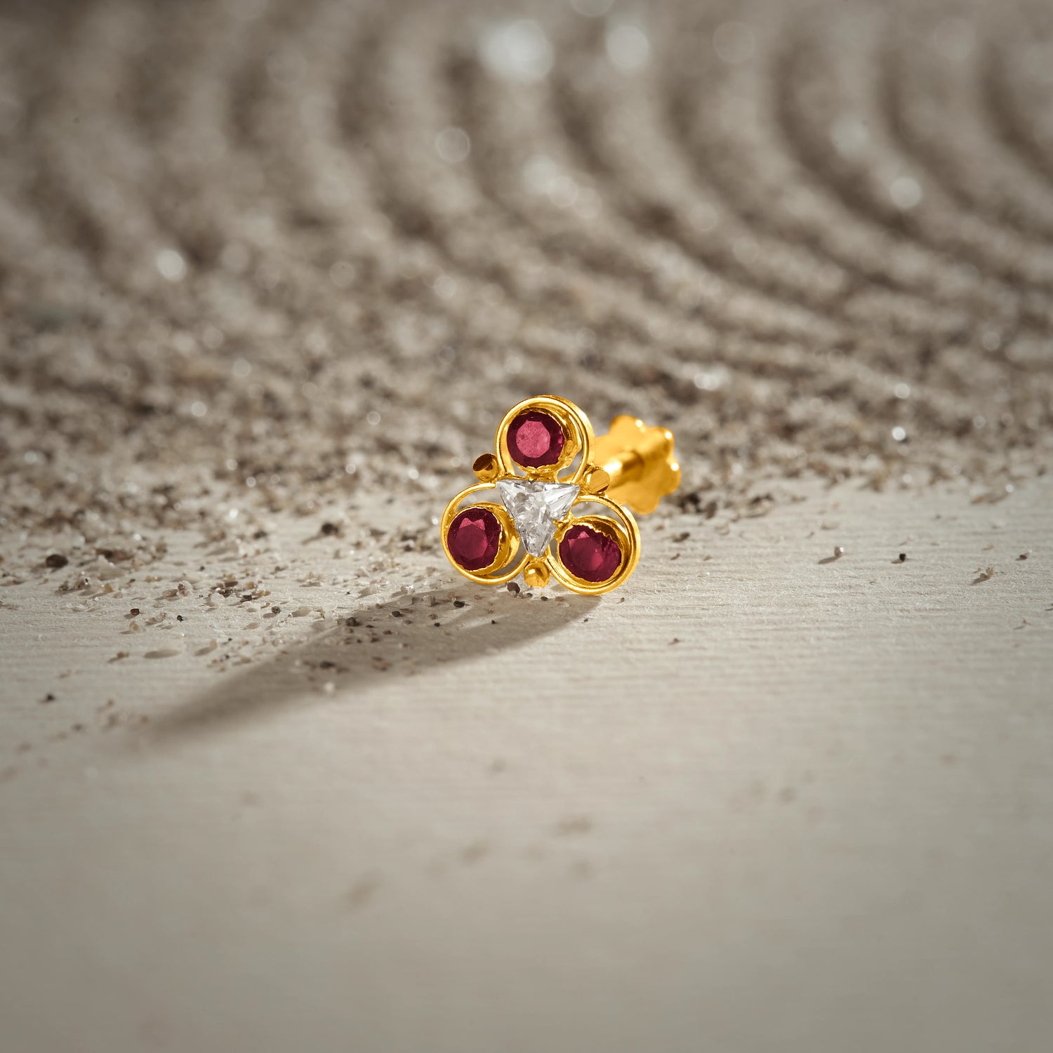 A gold trefoil-shaped stud earring with three ruby stones and a triangular white center stone, placed on a textured beige fabric background