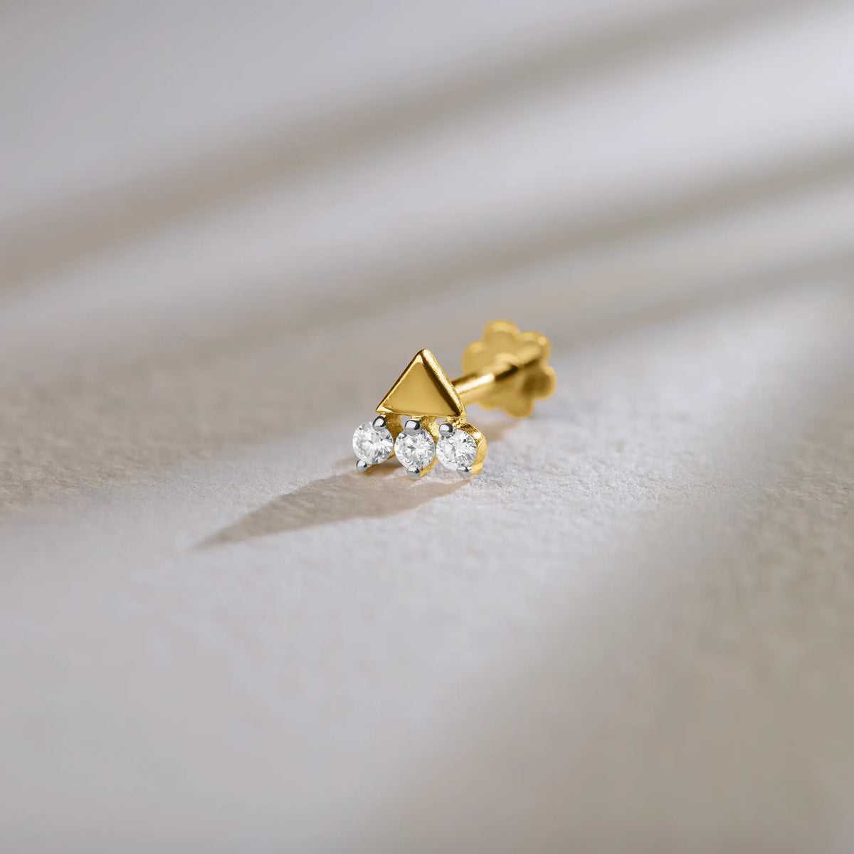 Product shot of a gold nose stud with a solid gold triangle above three gemstones