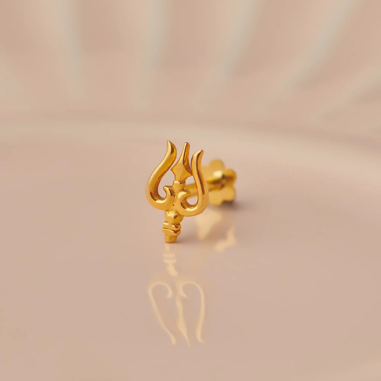 Gold trishul trident-shaped nose stud on beige background