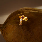 A gold vertical stud earring with a ruby stone at the top and three graduated white stones below, placed on a textured brown fabric background