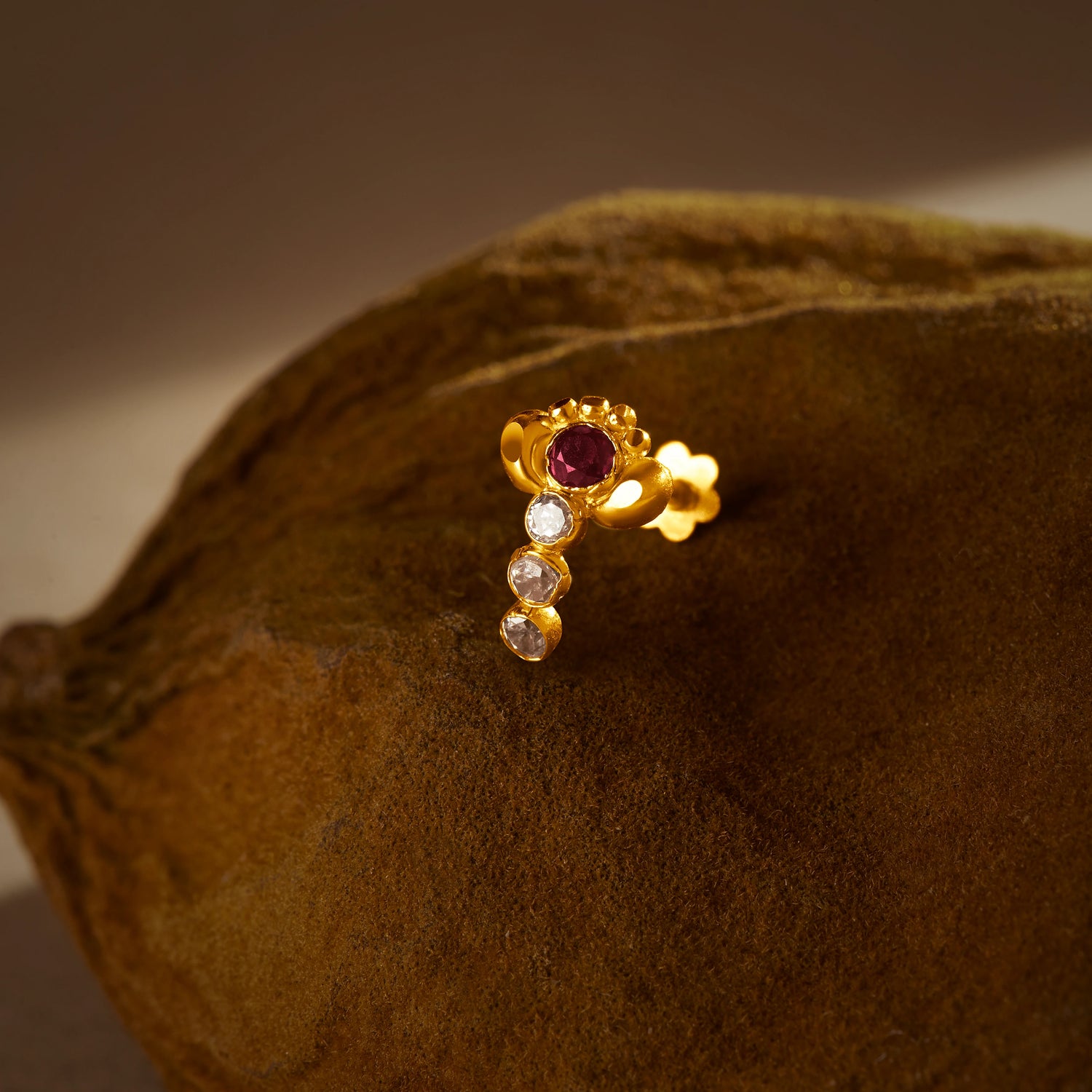A gold vertical stud earring with a ruby stone at the top and three graduated white stones below, placed on a textured brown fabric background