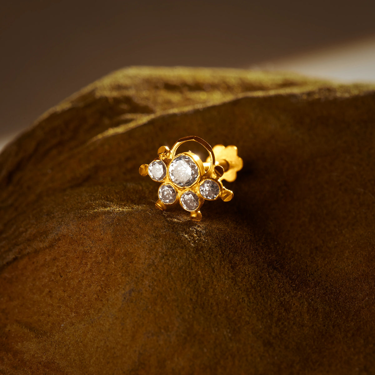 Close-up of a gold nose pin with a curved loop accent and a cluster of sparkling clear stones, set against a dark velvet backdrop