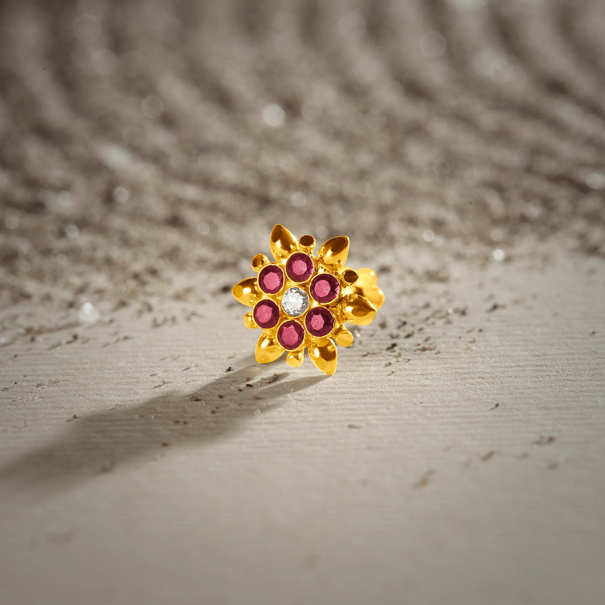 An 18K gold nose pin shaped like a starburst flower with pointed gold petals, set with ruby-red stones and a clear crystal center, photographed on a light textured surface