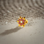 An 18K gold nose pin shaped like a starburst flower with pointed gold petals, set with ruby-red stones and a clear crystal center, photographed on a light textured surface