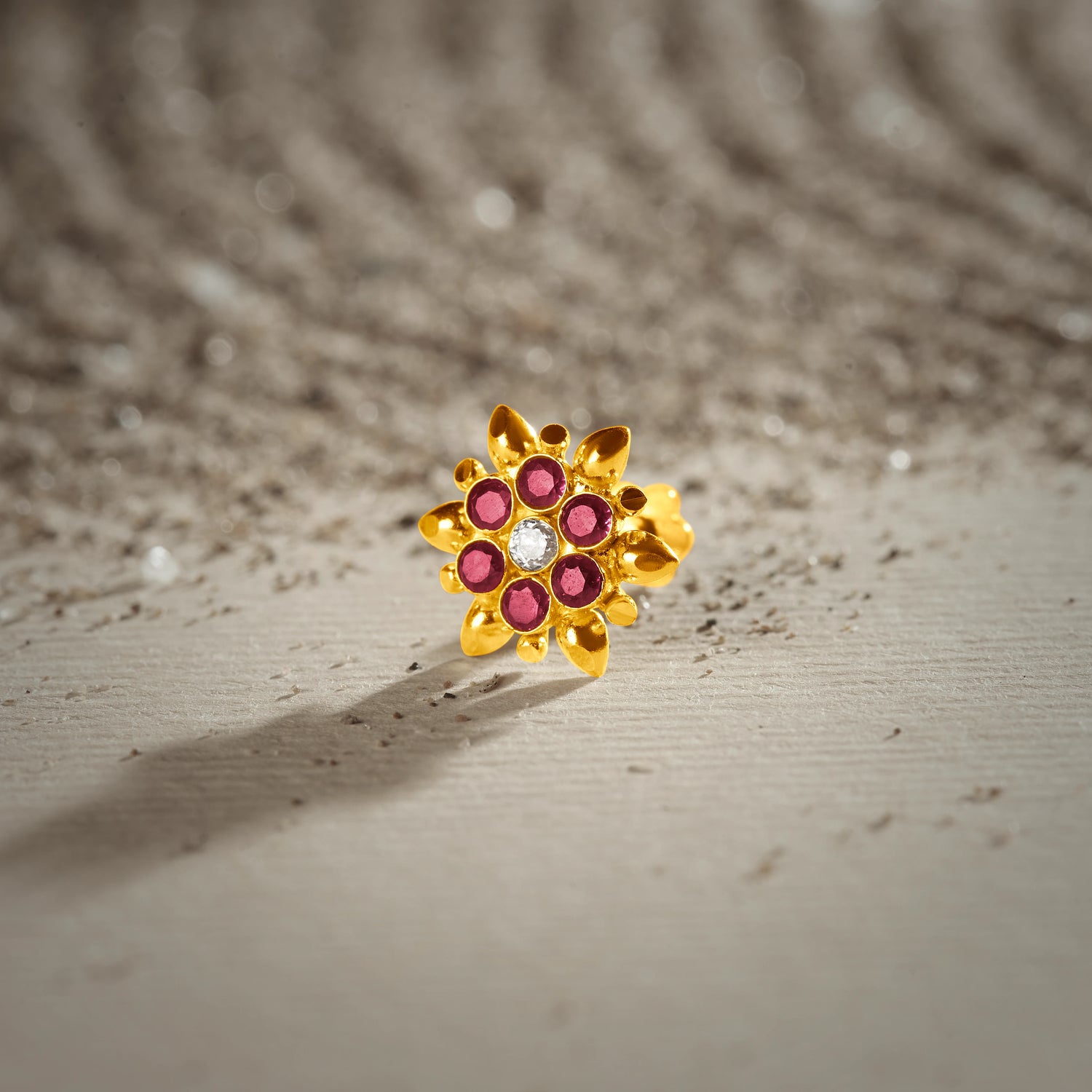 An 18K gold nose pin shaped like a starburst flower with pointed gold petals, set with ruby-red stones and a clear crystal center, photographed on a light textured surface