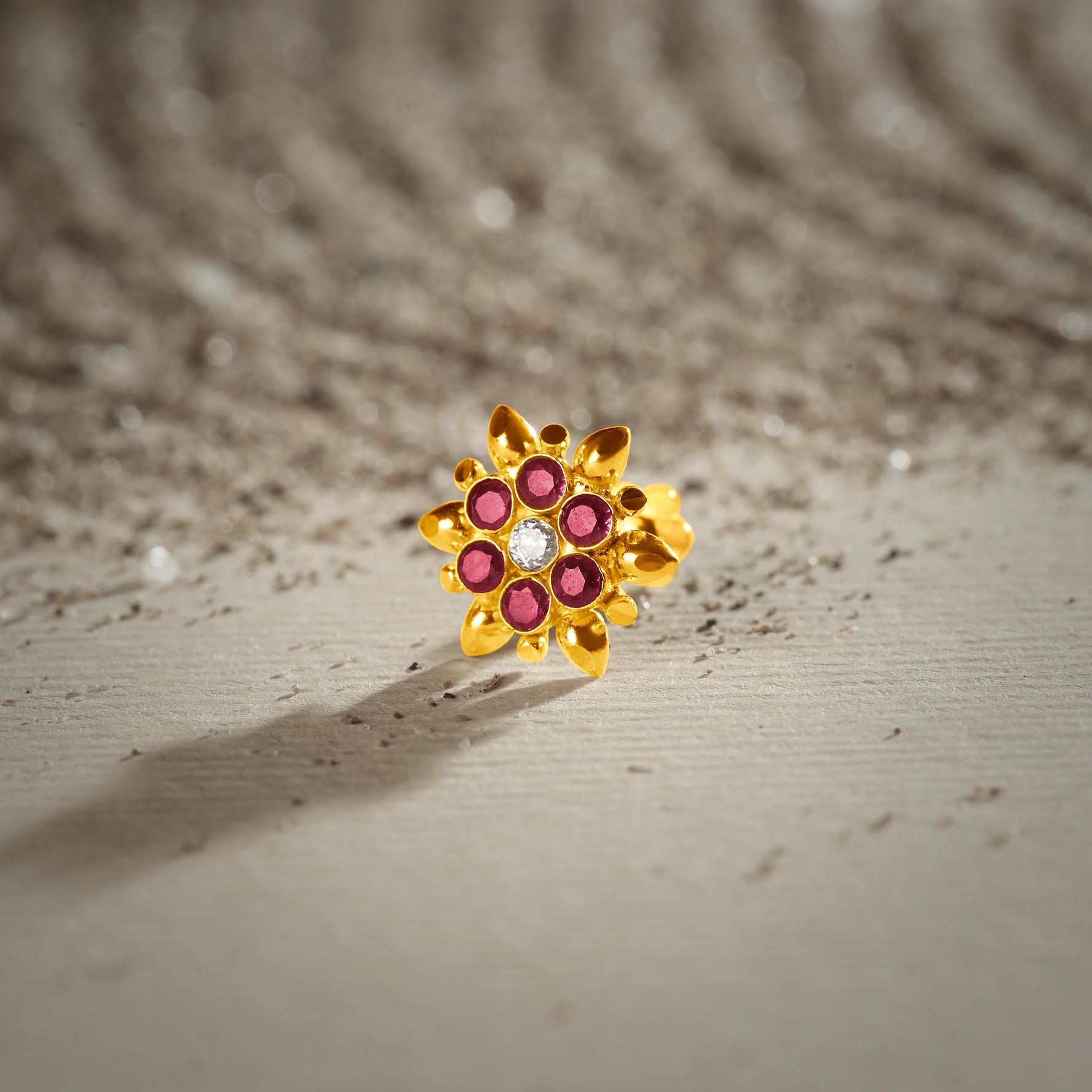 An 18K gold nose pin shaped like a starburst flower with pointed gold petals, set with ruby-red stones and a clear crystal center, photographed on a light textured surface