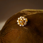 An 18K gold nose pin shaped like a dense two-row flower cluster, set with clear crystal stones, photographed on a dark textured fabric background