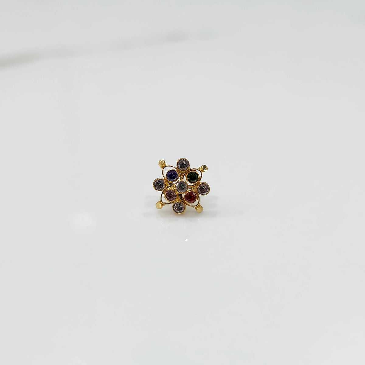 A gold nose pin shaped like a flower, set with multicolored gemstones in shades of red, blue, green, and purple