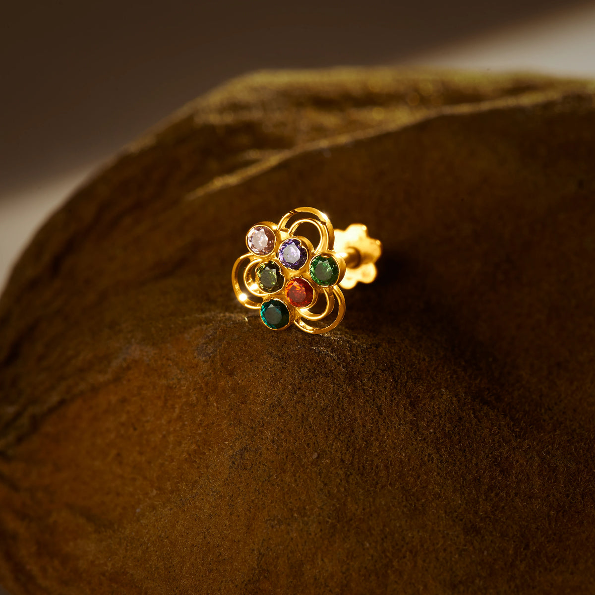 A gold nose pin shaped like a flower with open loop petals, set with multicolored stones in purple, green, and red, photographed on a dark textured fabric background