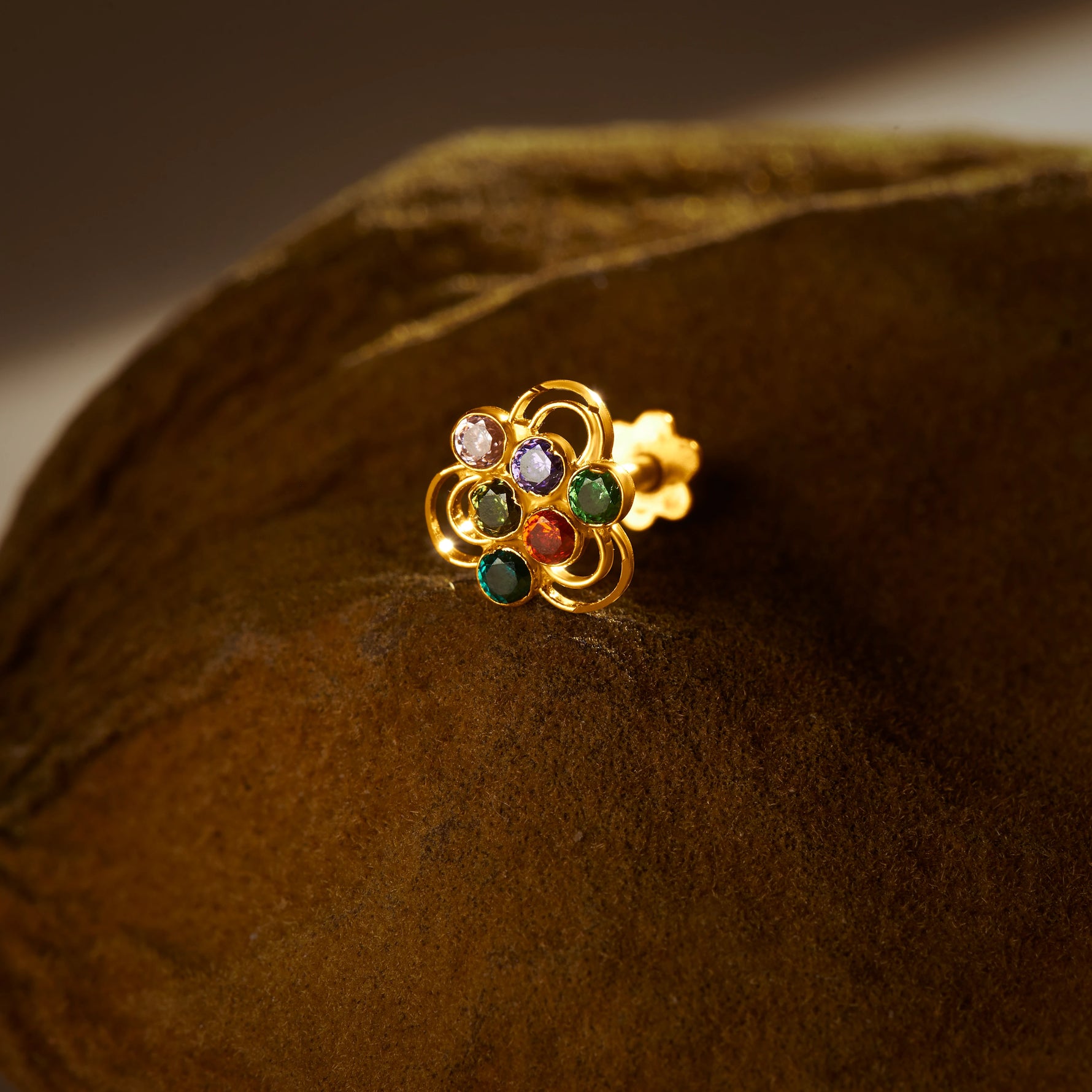 A gold nose pin shaped like a flower with open loop petals, set with multicolored stones in purple, green, and red, photographed on a dark textured fabric background
