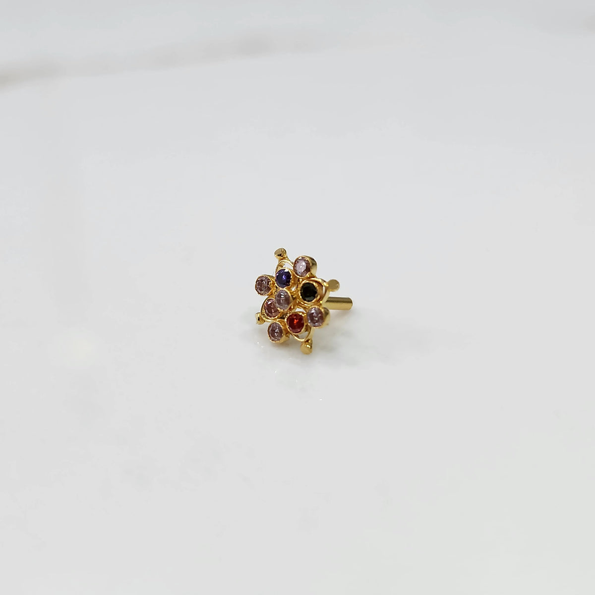 A gold nose pin shaped like a flower, set with multicolored gemstones including red, blue, and champagne tones