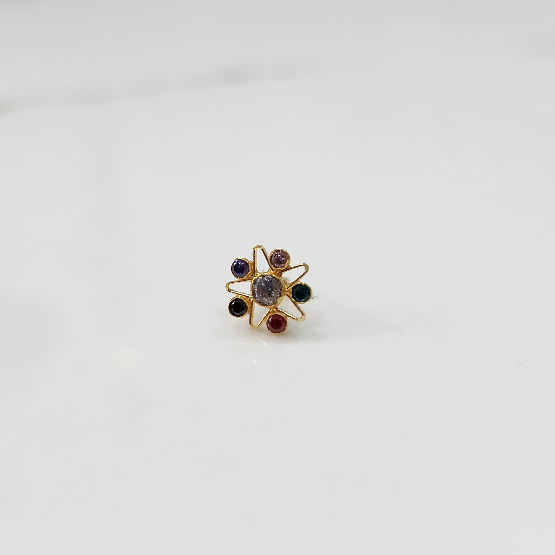 A gold nose pin shaped like a star with a pale grey center stone and multicolored accent stones in purple, pink, green, black, and red