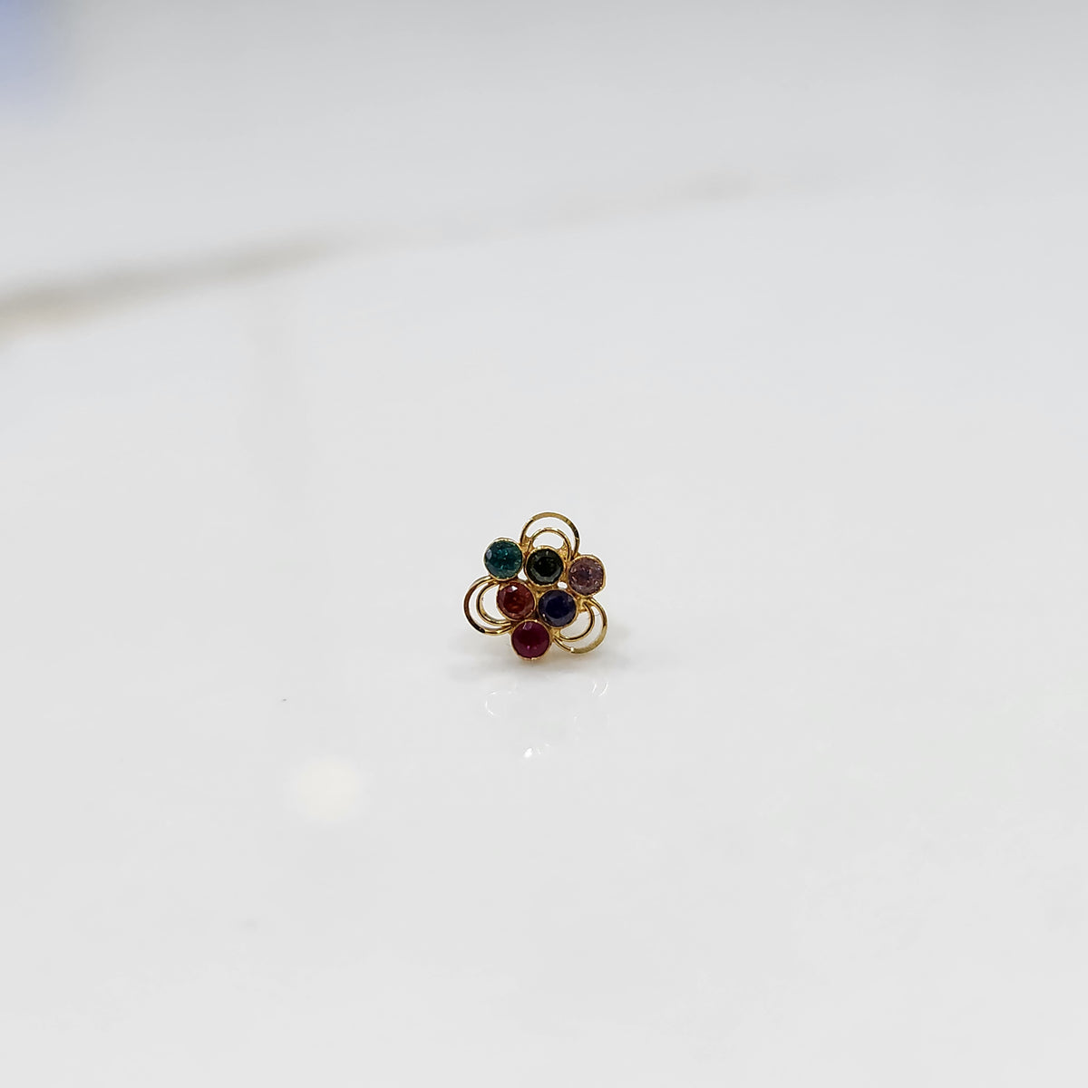 An 18K gold nose pin shaped like a flower, set with multicolored stones in teal, purple, red, and blue, each in an open gold loop