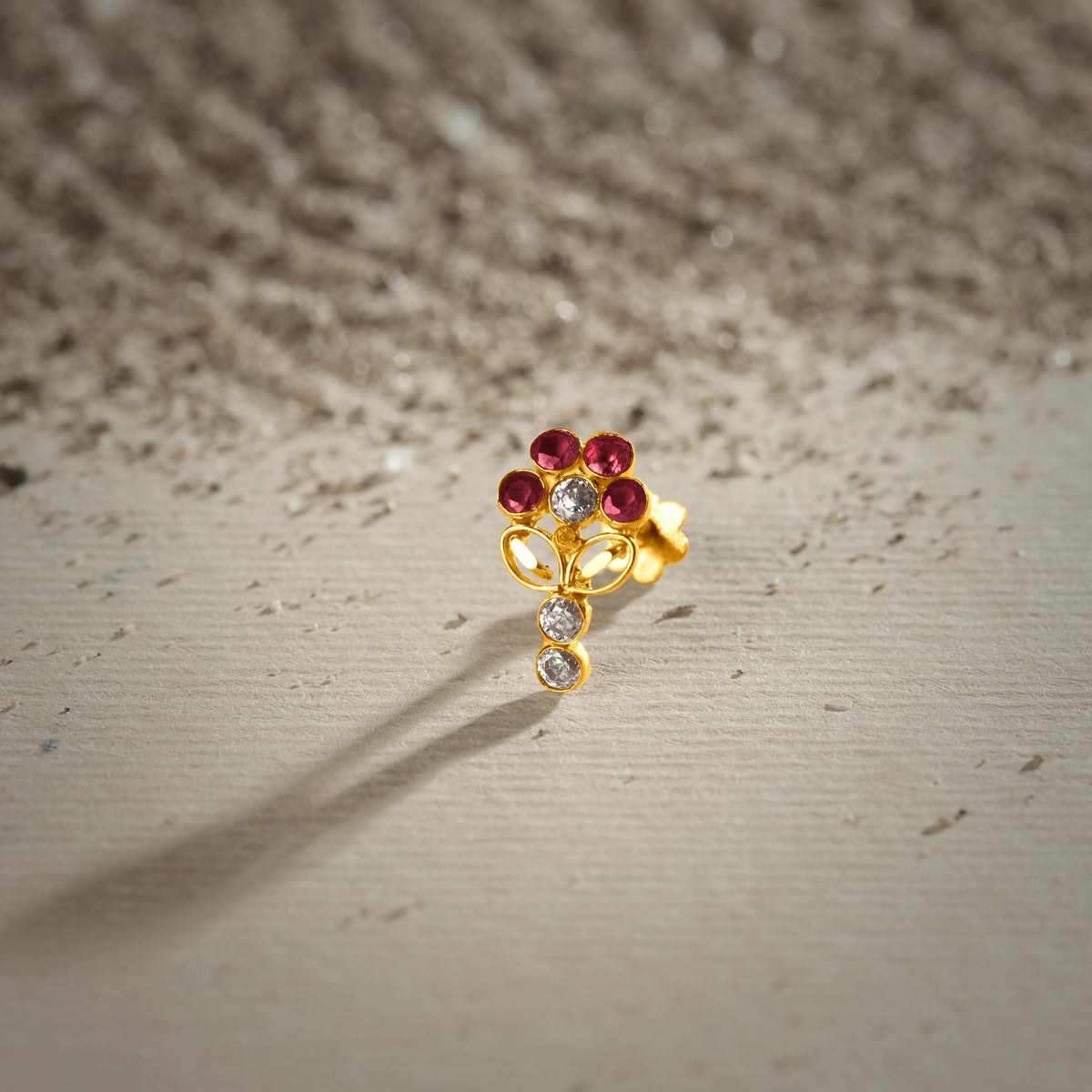 An 18K gold nose pin shaped like a flower with ruby-red petals and a clear crystal center, with two hanging clear crystal drops below, photographed on a light textured surface
